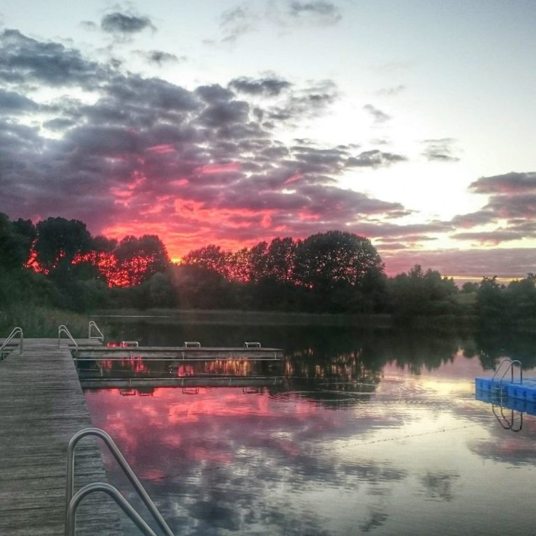 Unser Freibad e.V. Grevesmühlen Sonnenuntergang über einem ruhigen Teich, Spiegelung von Bäumen und Wolken im Wasser.