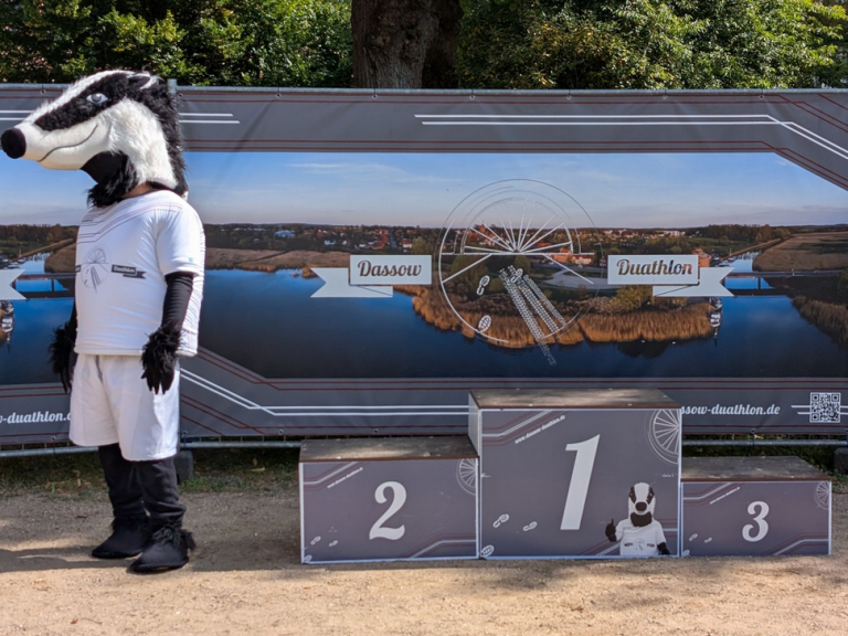 Dassow Duathlon 2026 Maskottchen im Tierkostüm steht neben einem Präsentationsbanner und Medaillen auf einem Tisch.